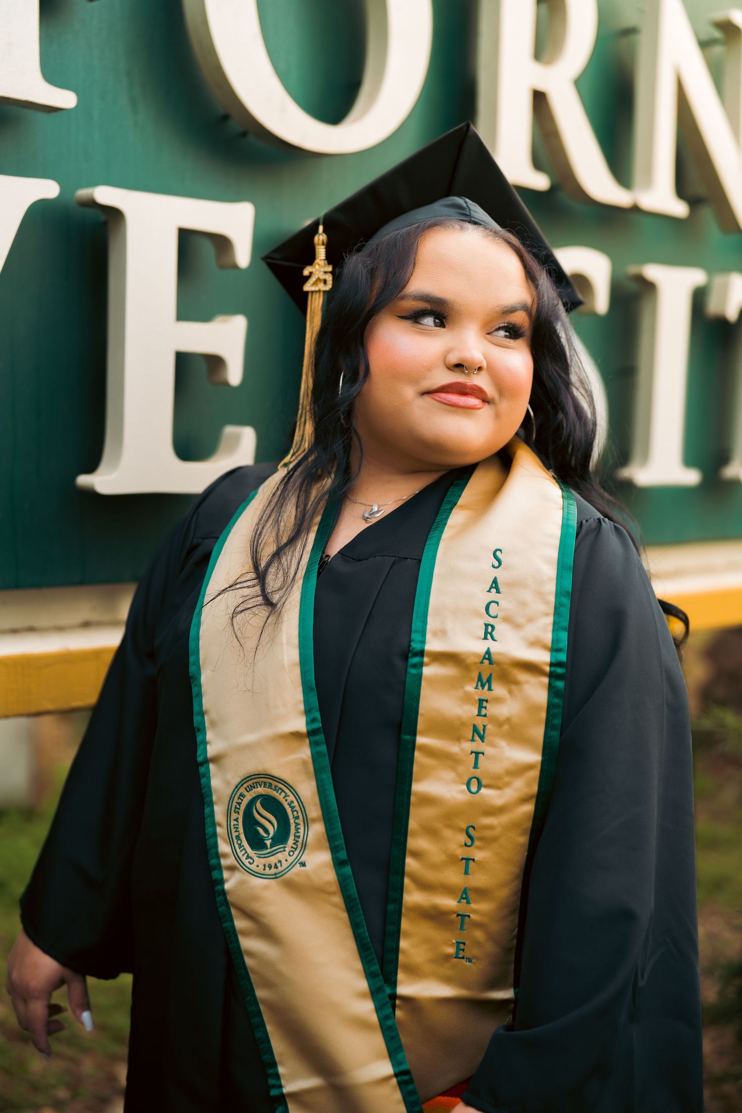 Smiling graduate in black cap and gown wearing Sacramento State stole standing near green university sign.