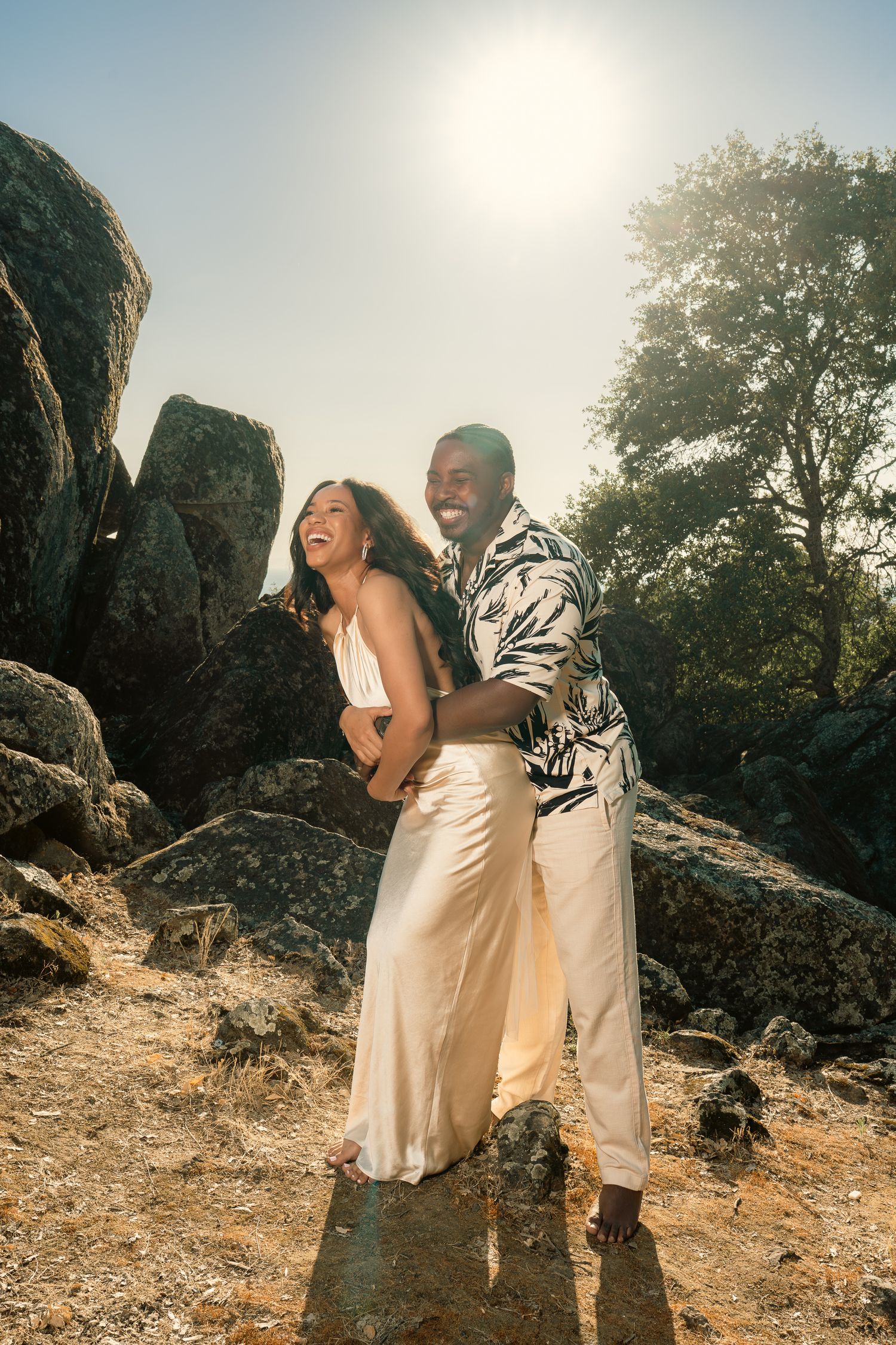 Joyful newlyweds laughing and embracing among rocky terrain in bright sunlight.