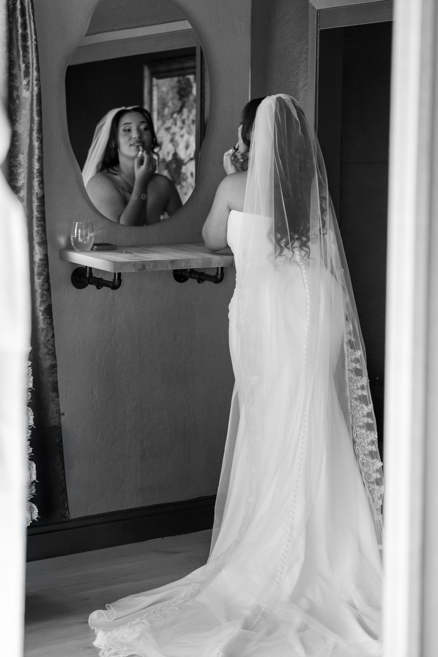 Black and white photo of bride in lace veil applying makeup while looking in mirror before wedding.