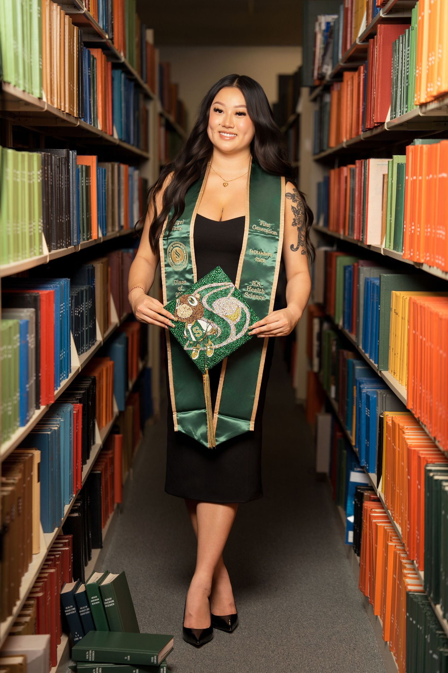 Smiling graduate in black dress with green stole holding decorated cap in a library aisle.