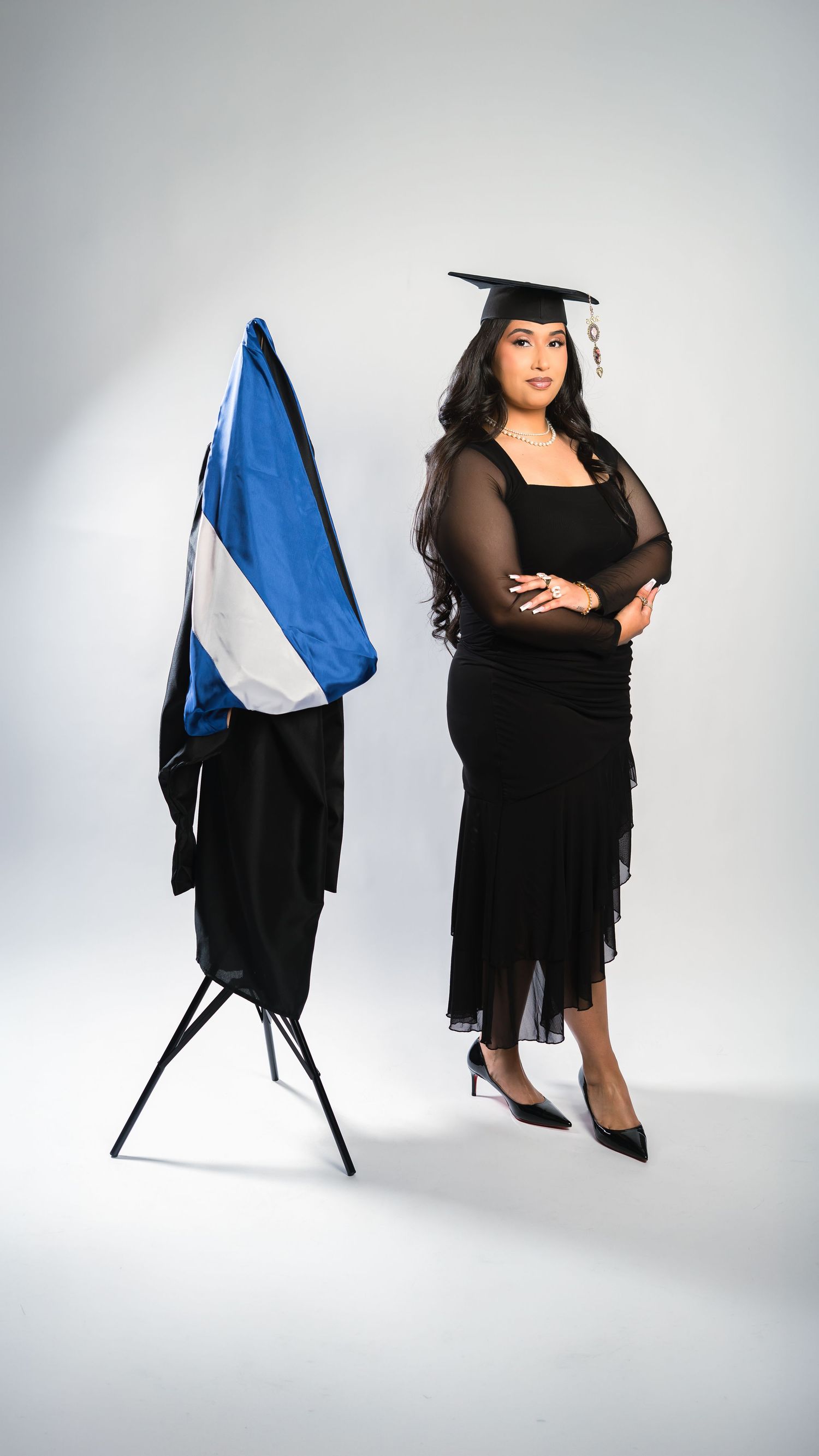 Female graduate in black dress standing beside blue and white flag with cap.