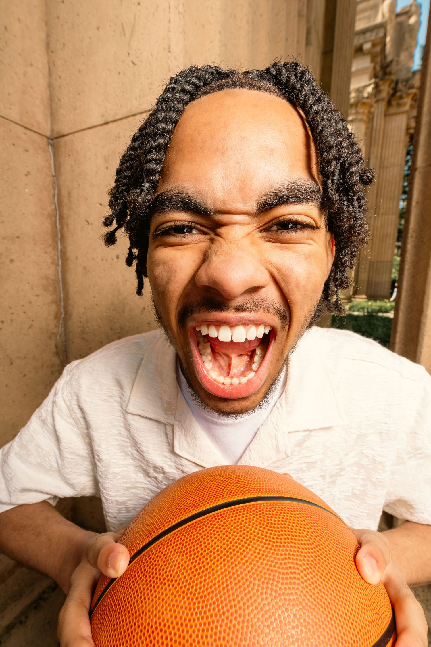 Energetic young man screaming while holding basketball close to camera at stone column backdrop.