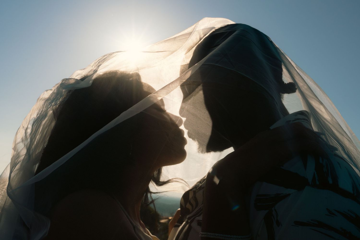 Bride and groom sharing an intimate moment under a wedding veil backlit by golden sunlight.