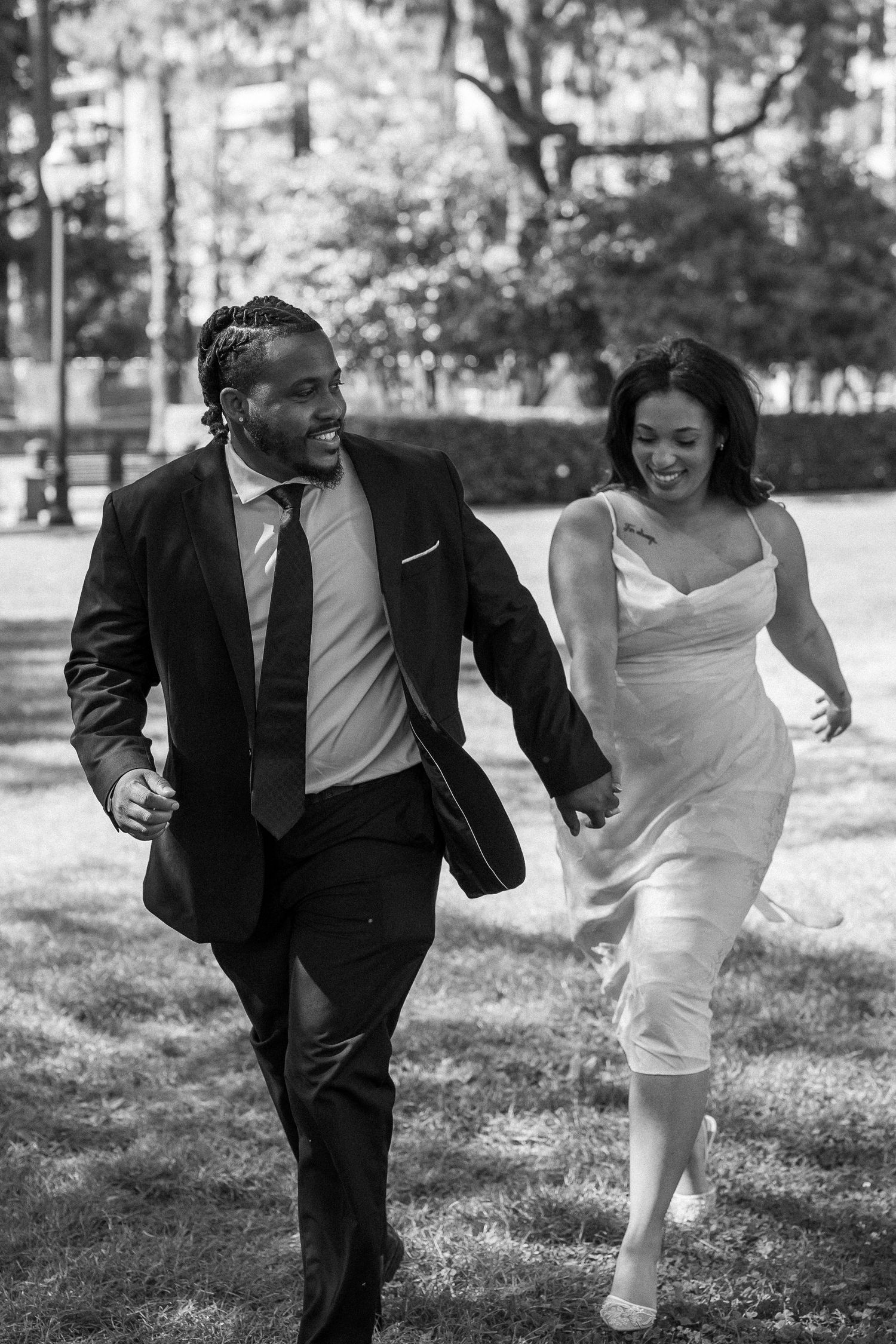 Happy newlywed couple running hand-in-hand through a park, bride in white dress and groom in black suit.
