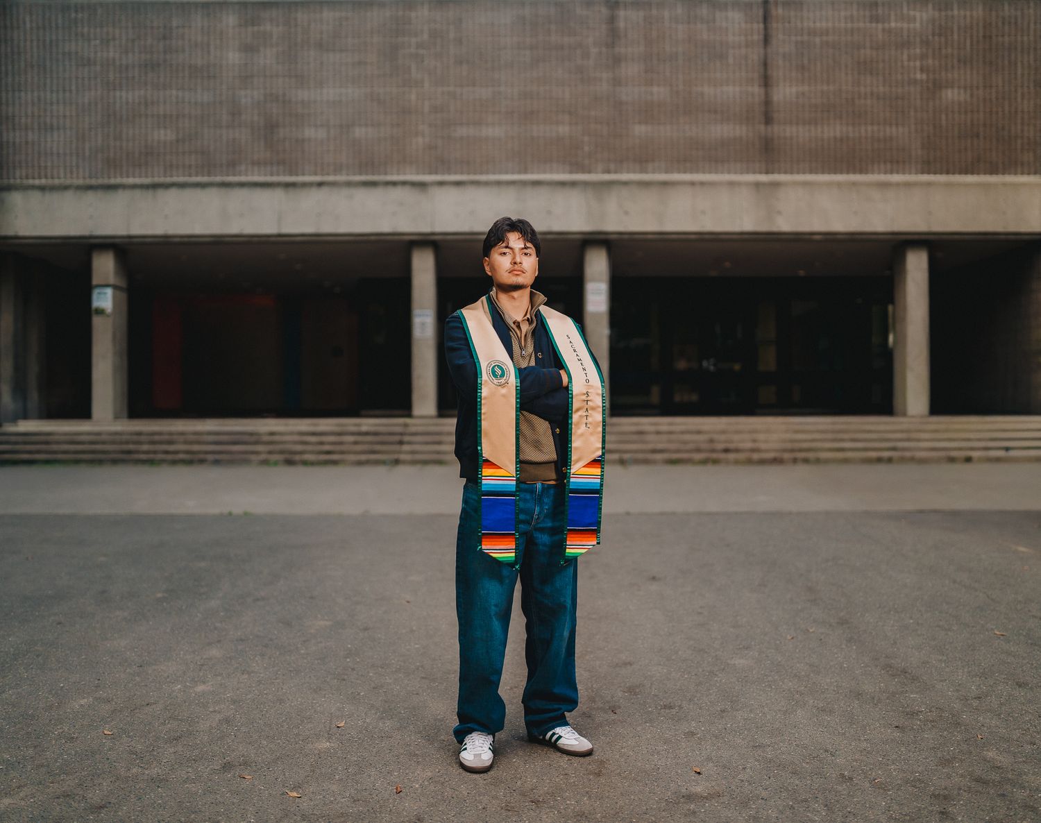 Graduate student stands proudly wearing colorful ceremonial stole outside a university building.