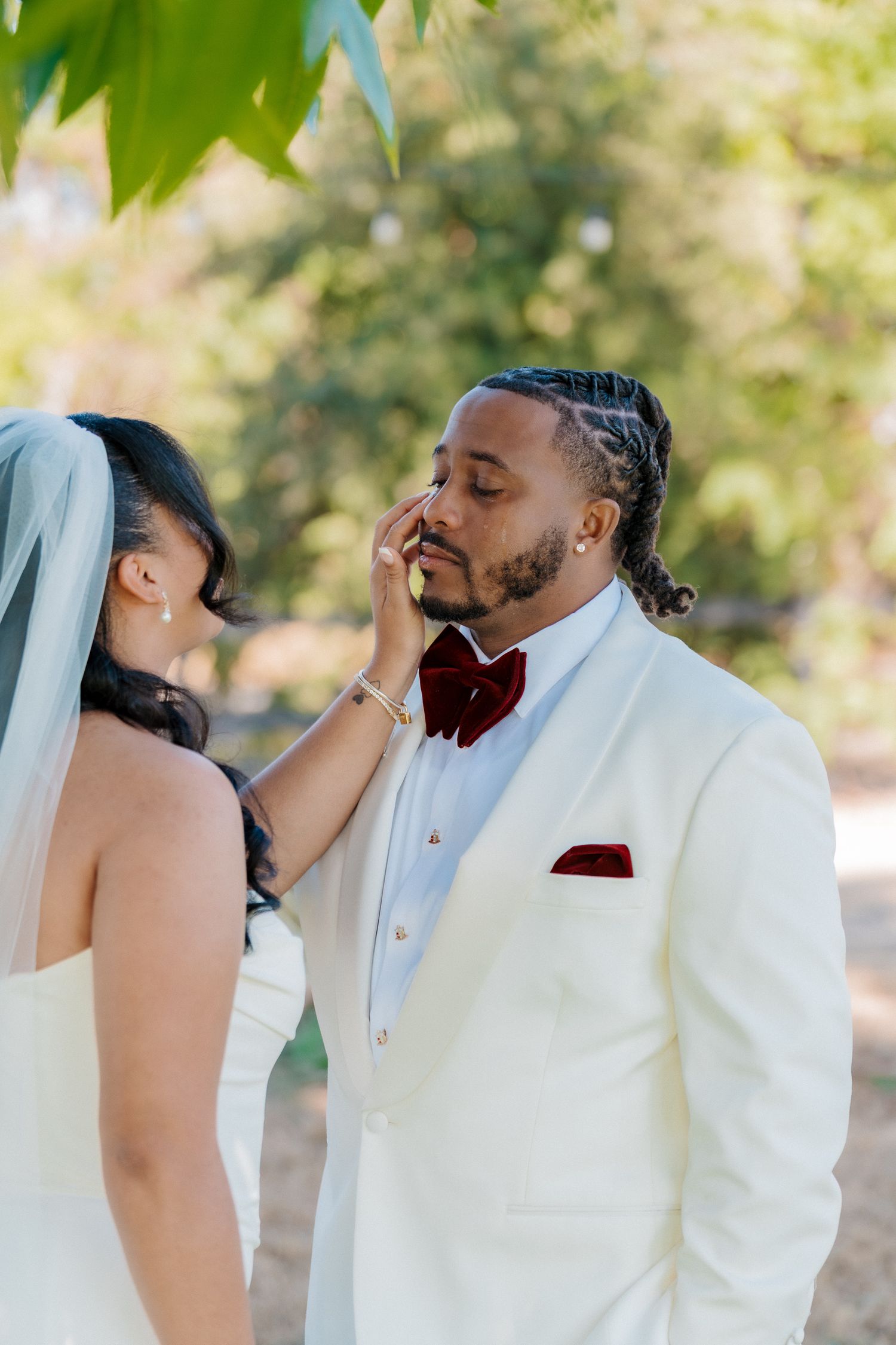 Bride gently touches grooms face as he tears up in white tuxedo with burgundy bow tie.