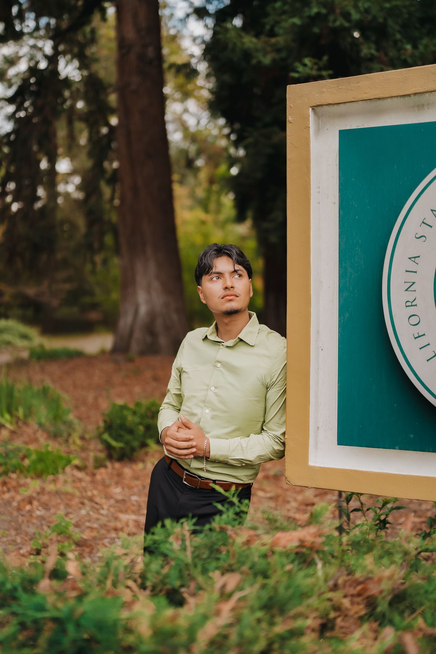 Young man in green shirt poses thoughtfully beside a California State University sign surrounded by lush forest.