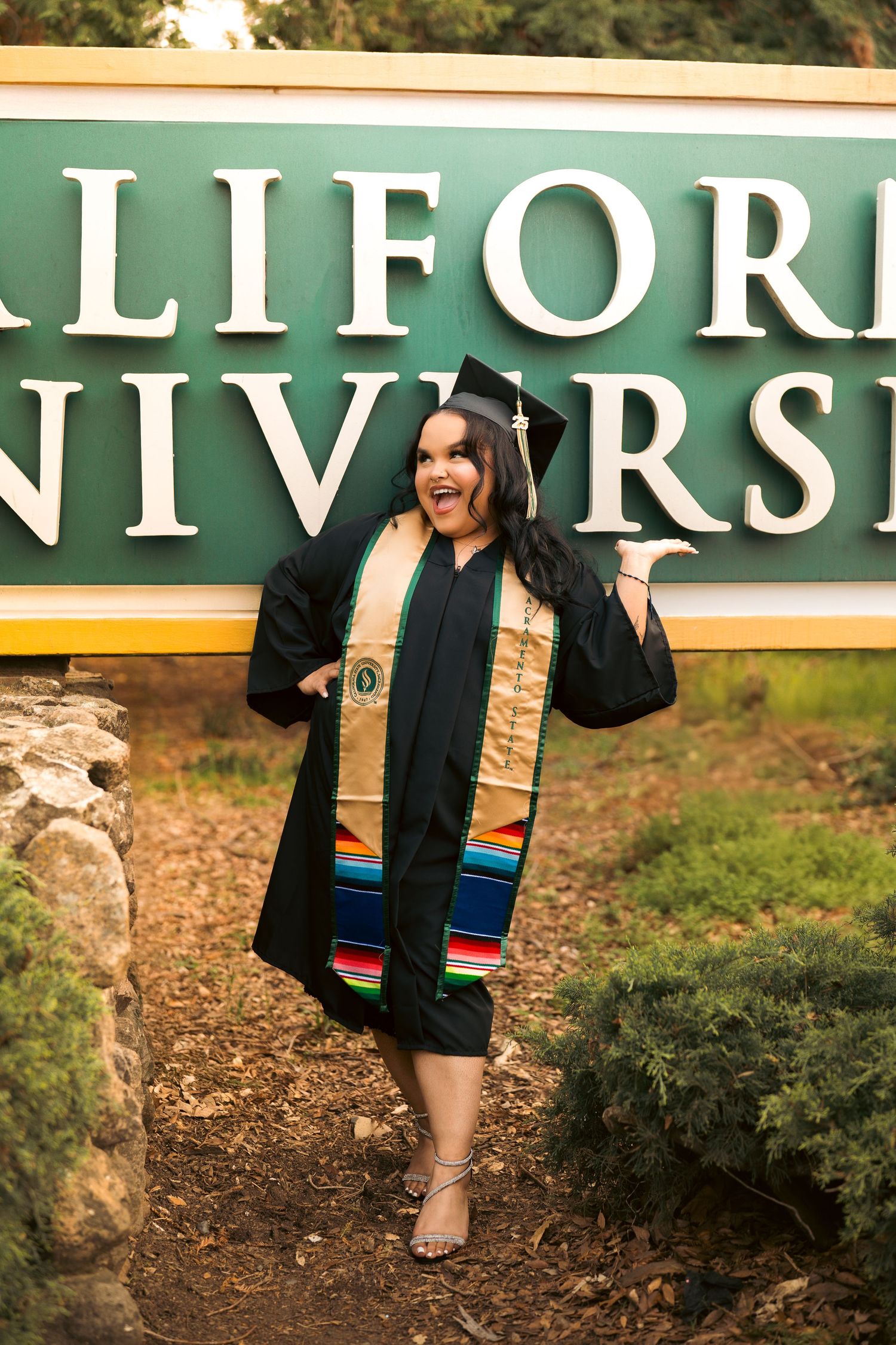 Joyful graduate in black cap and gown with colorful stole posing by California University green sign.