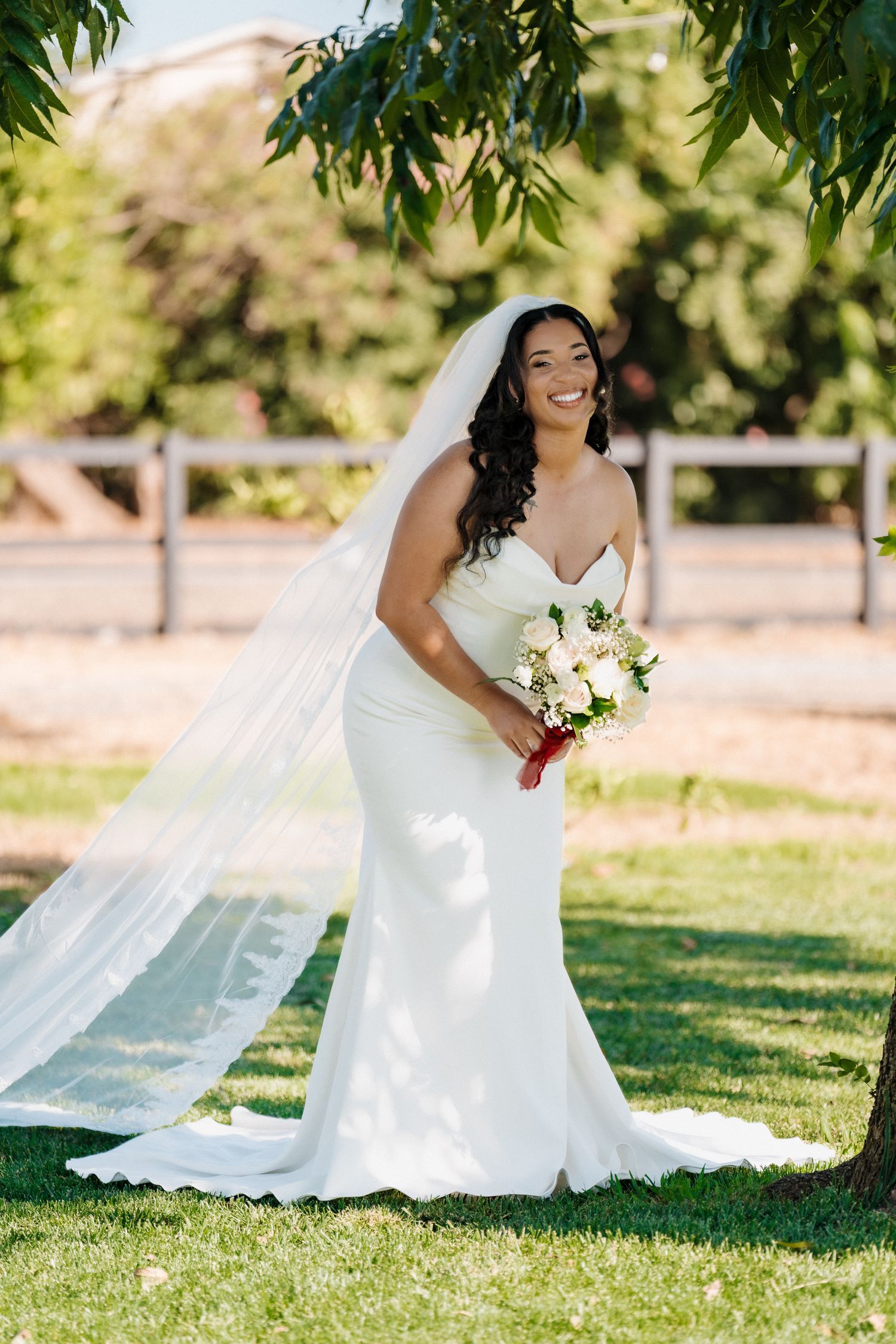 Smiling bride in white mermaid gown and veil holds bouquet outdoors on green lawn.
