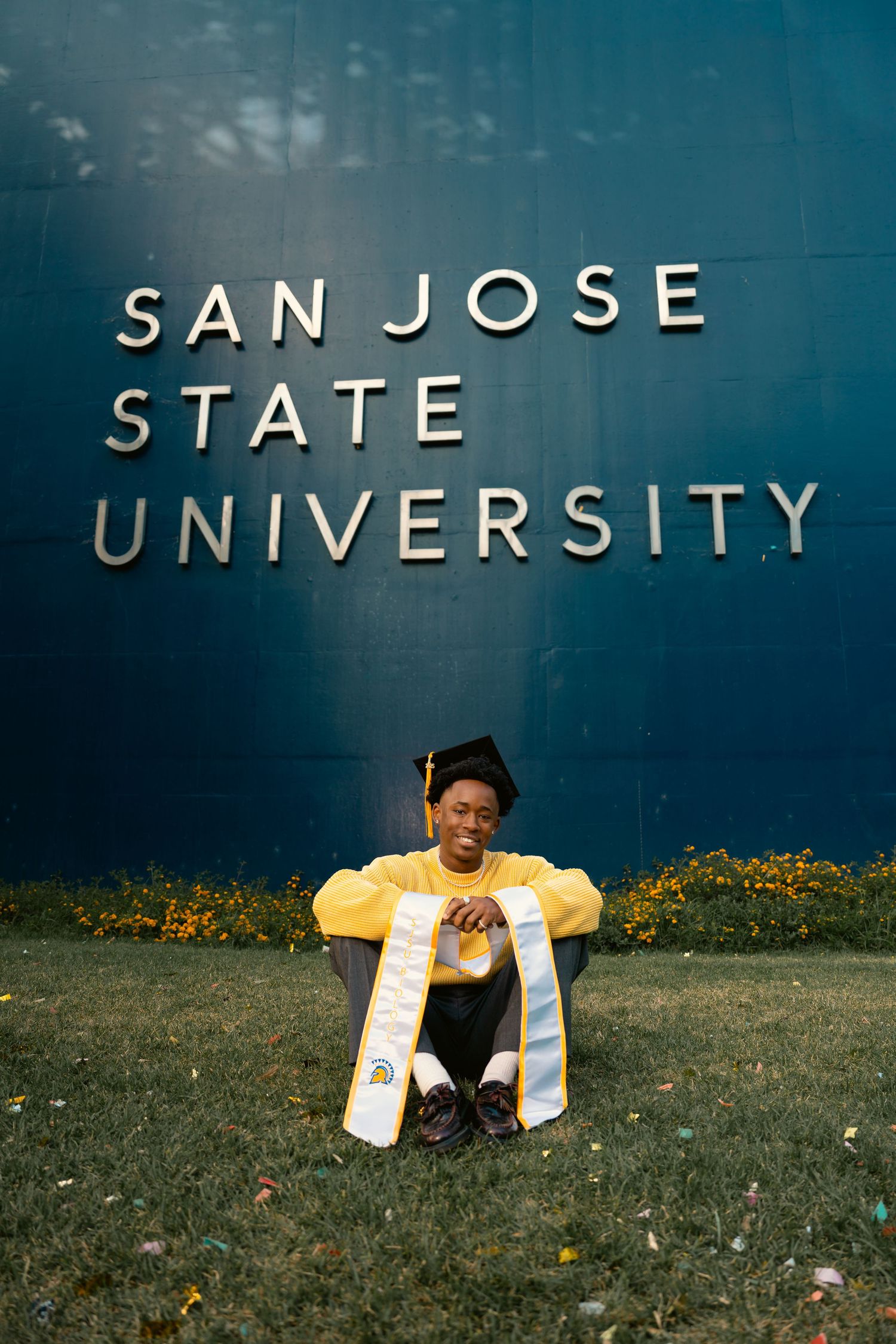 Graduate in cap and yellow sweater sitting on grass in front of San Jose State University sign.