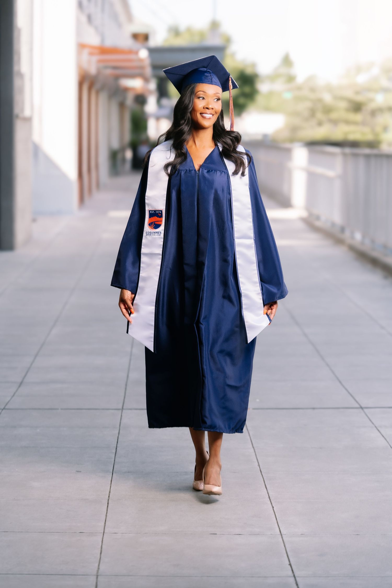 Smiling Black woman in full navy graduation cap and gown walking confidently down an outdoor corridor.