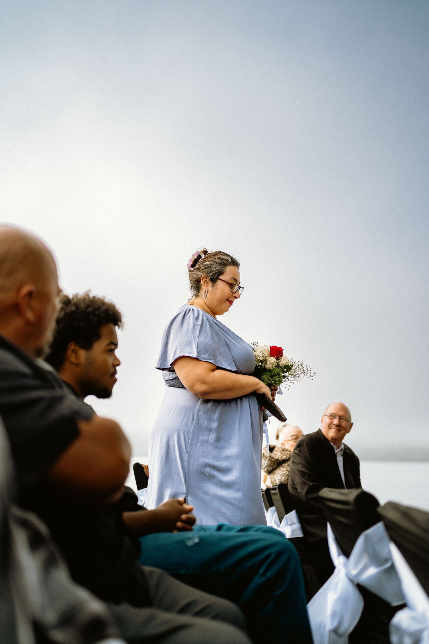 Woman in blue dress holding red roses stands among wedding guests seated outdoors near waterfront.