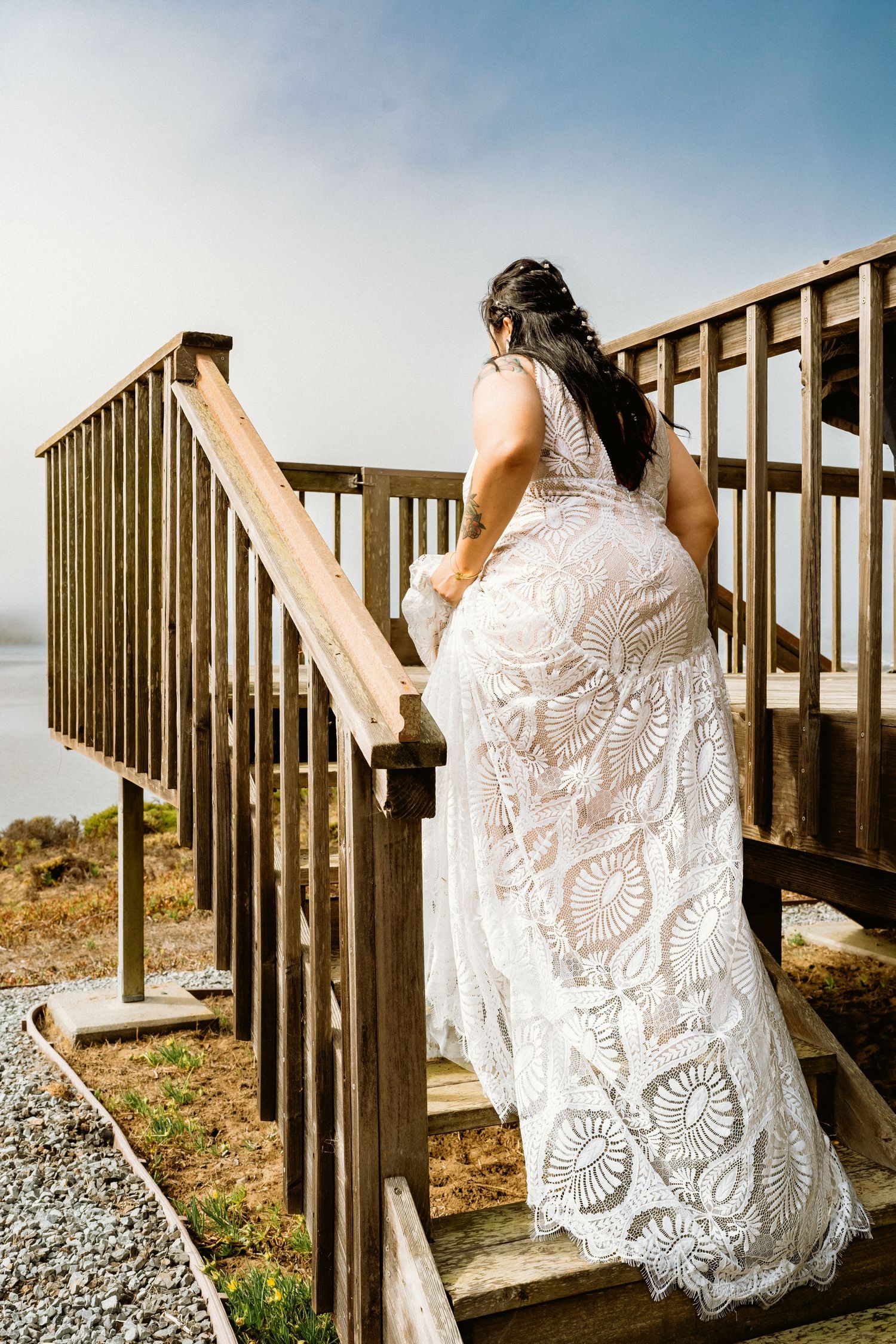 Bride in intricate white lace wedding gown ascends wooden stairs overlooking a scenic waterfront.