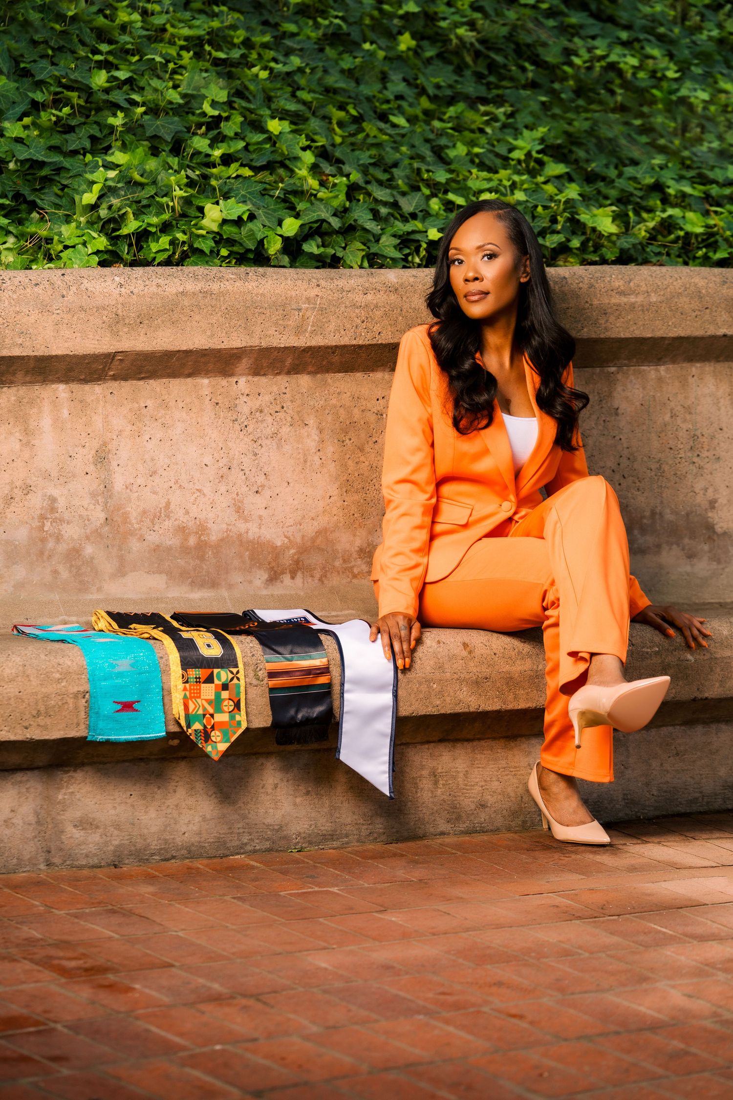 Elegant woman in orange pantsuit sitting on stone bench beside colorful kente cloth graduation stole.