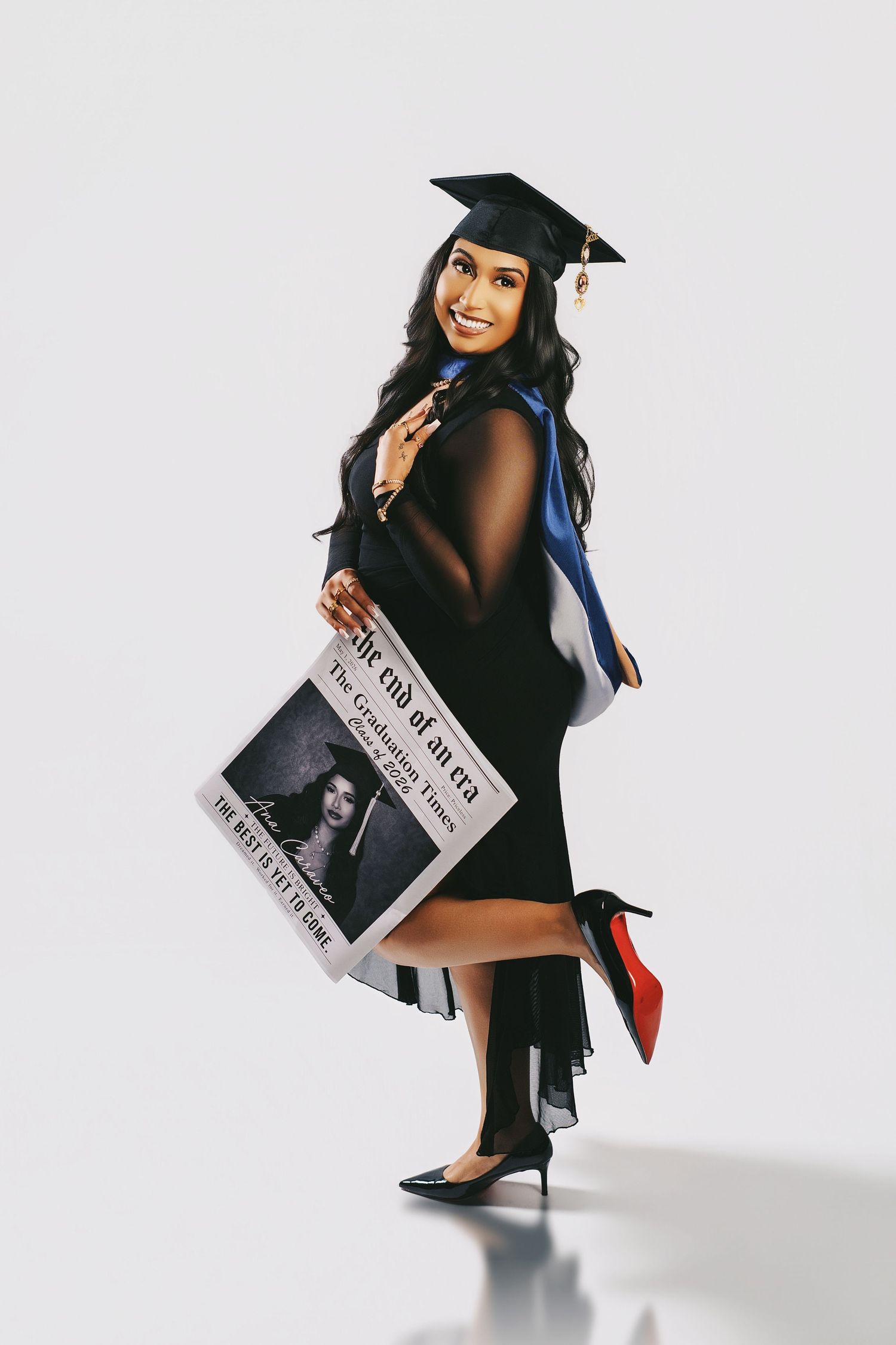 Smiling graduate in black cap and gown holding a decorative newspaper prop in a studio photo shoot.