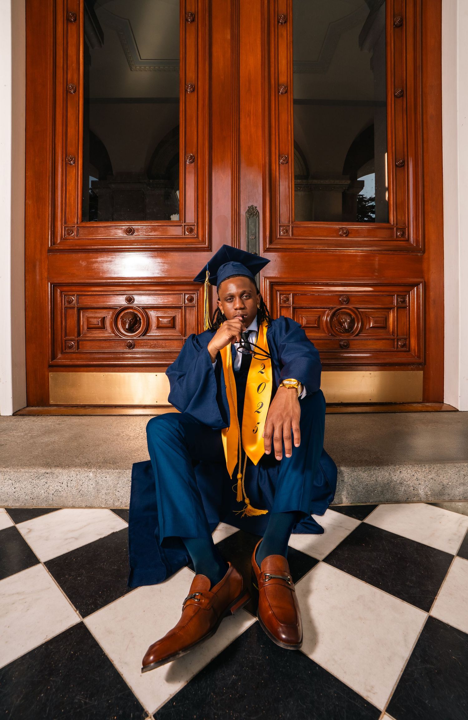 Graduate in blue cap and gown sitting on checkered floor before ornate wooden doors.