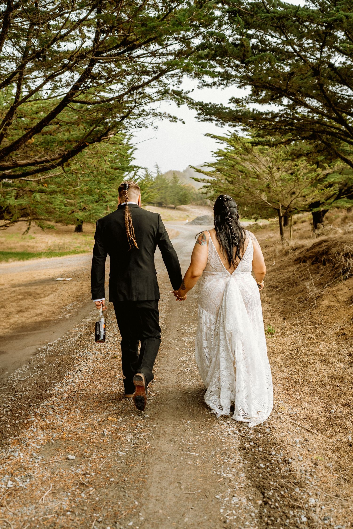 Newlywed couple walks hand in hand down tree-lined path after outdoor wedding ceremony.