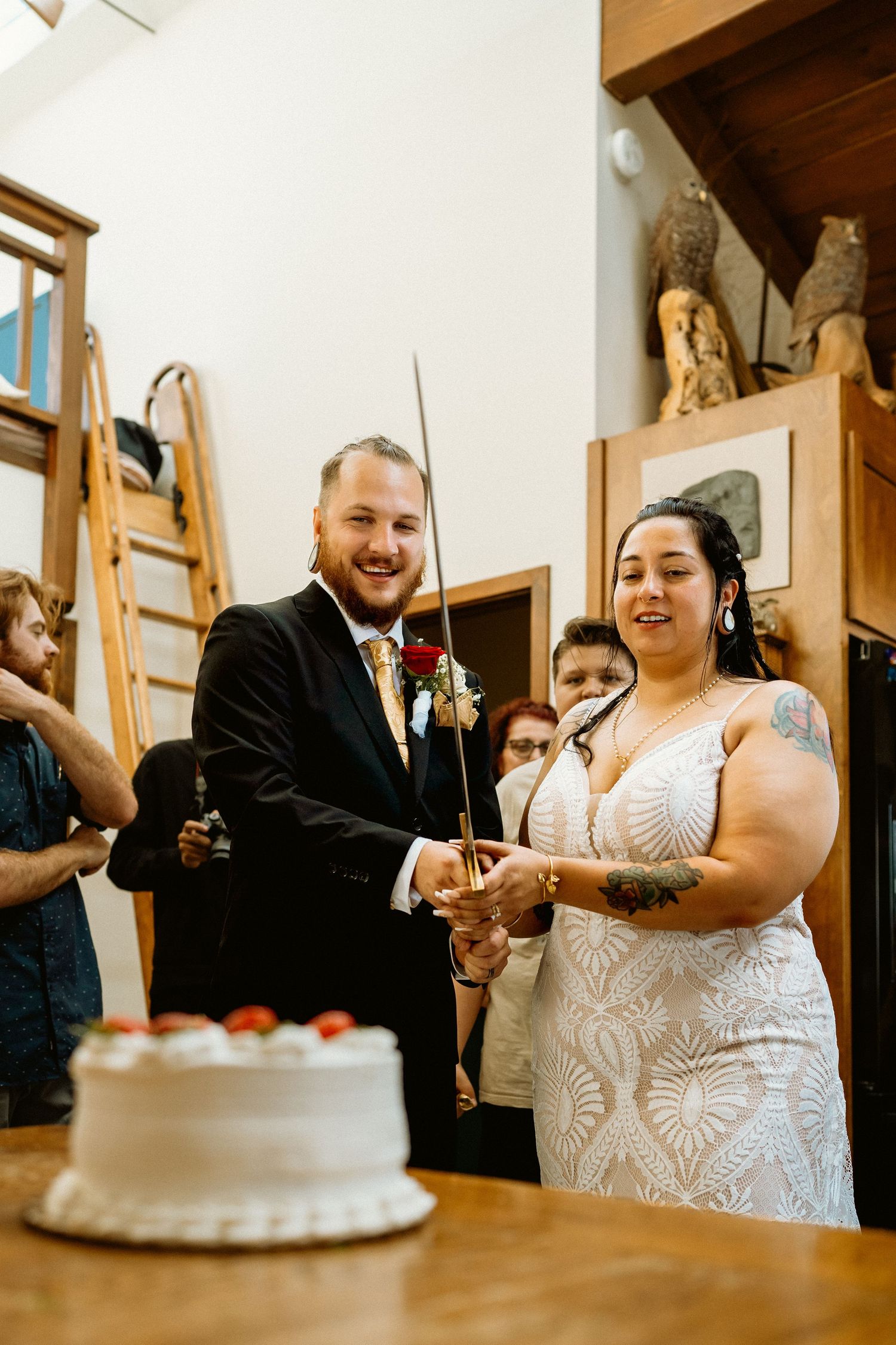 Smiling newlyweds cut wedding cake with a sword at rustic indoor wedding reception.