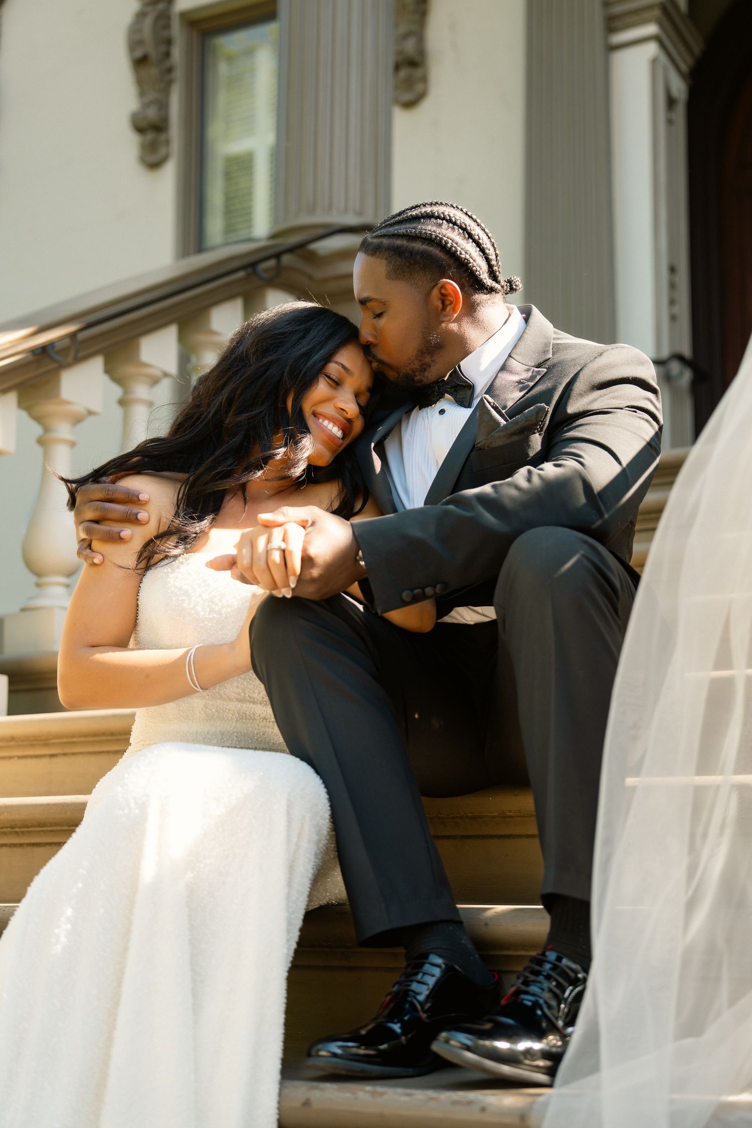 Groom kisses bride on steps of grand building while she laughs joyfully.
