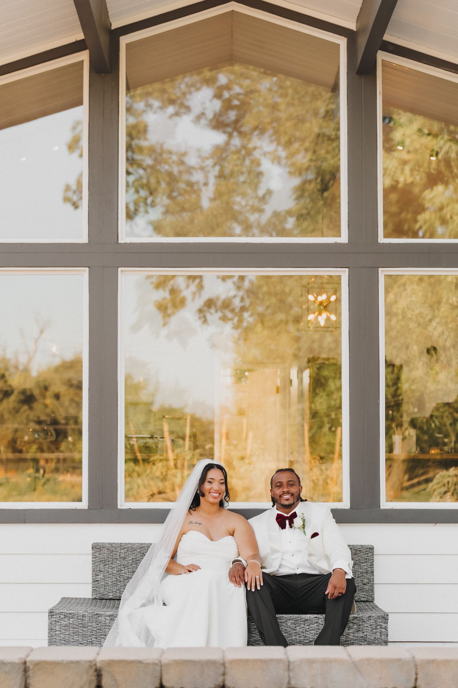 Smiling newlyweds sit together in front of a large chapel window in warm golden evening light.