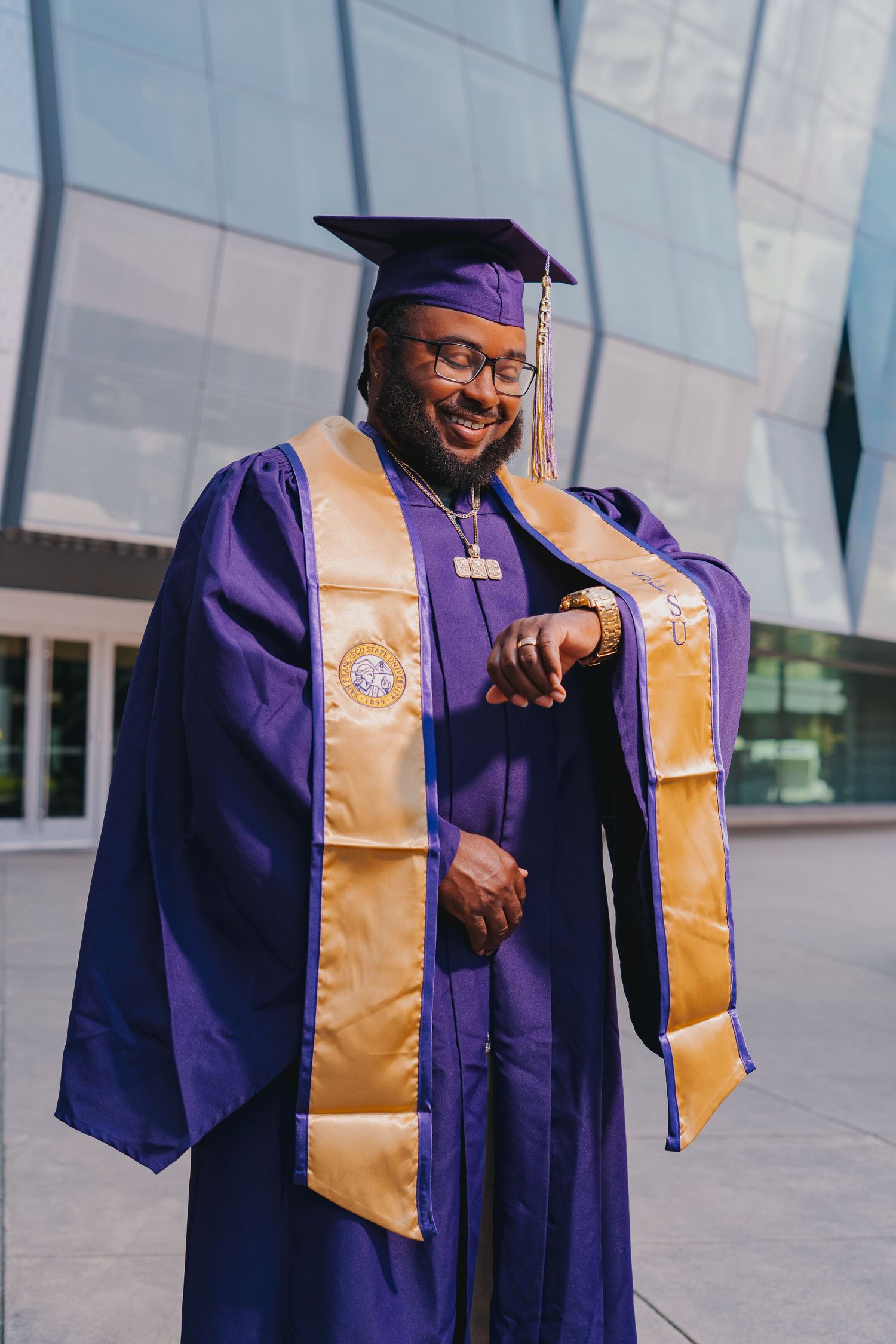 Graduate in purple and gold SFSU cap and gown smiles while checking watch outdoors near glass building.