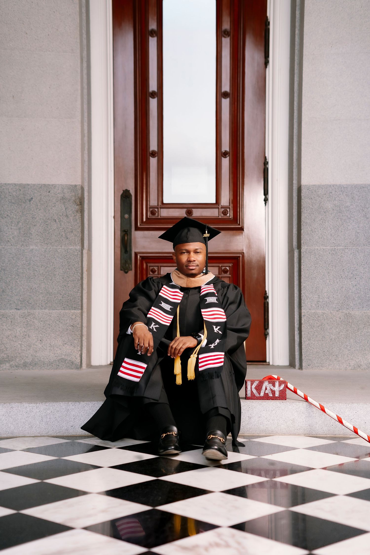 Graduate in cap and gown sitting on checkered floor holding small American flags.