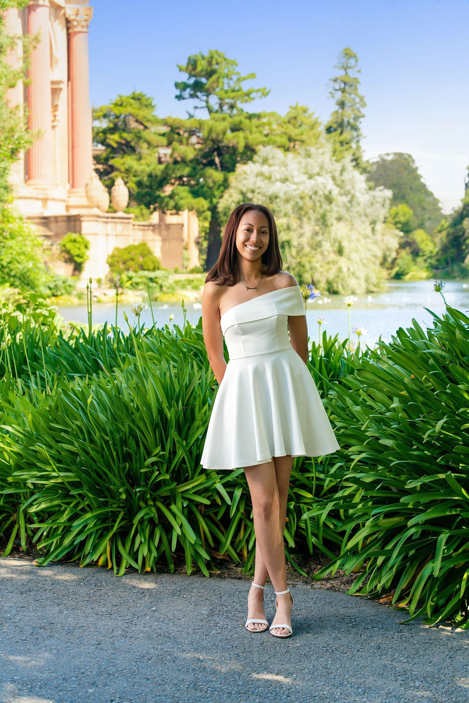 Smiling young woman in white off-shoulder mini dress posing at Palace of Fine Arts gardens.