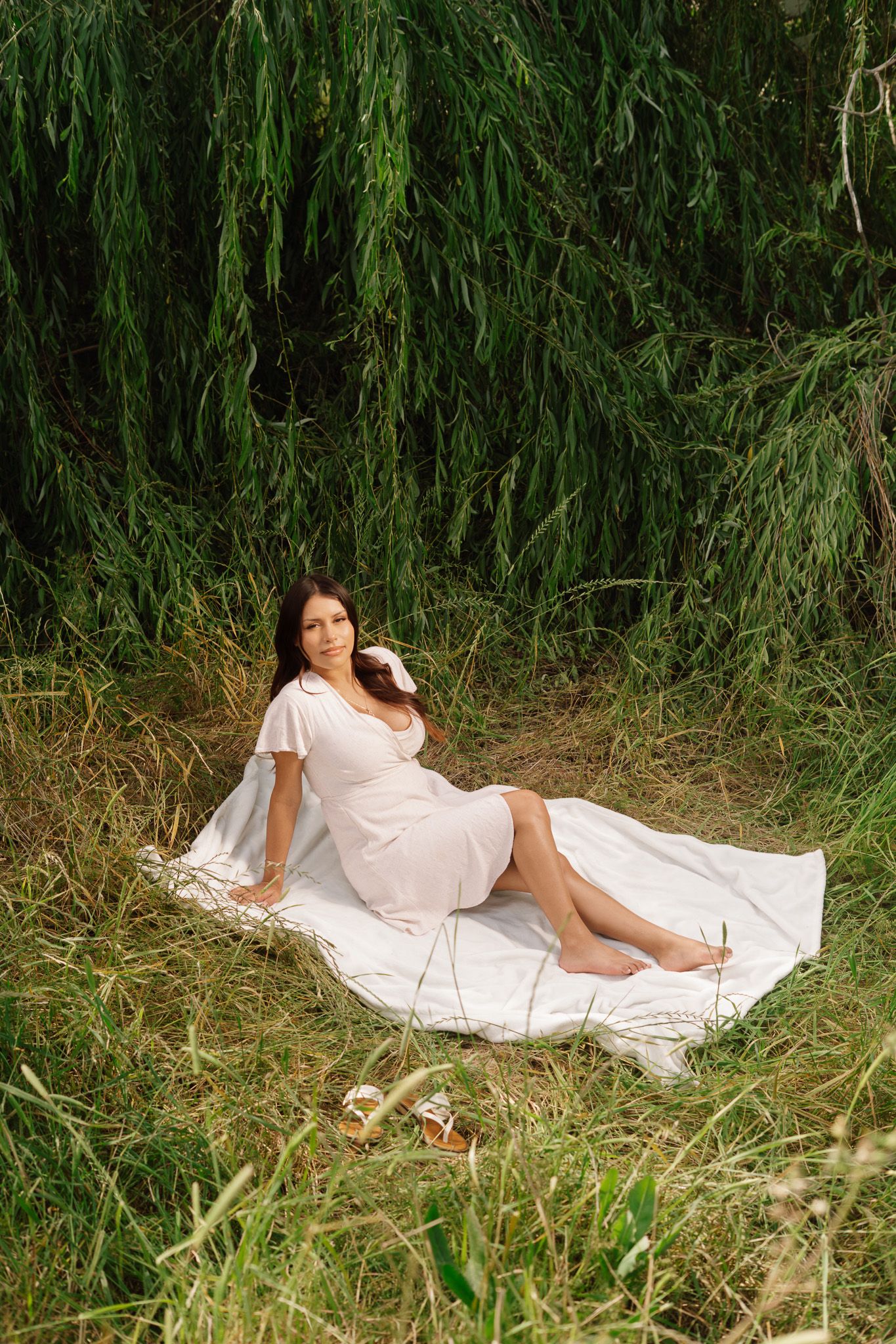Pregnant woman in white dress relaxing on blanket under weeping willow tree in nature.