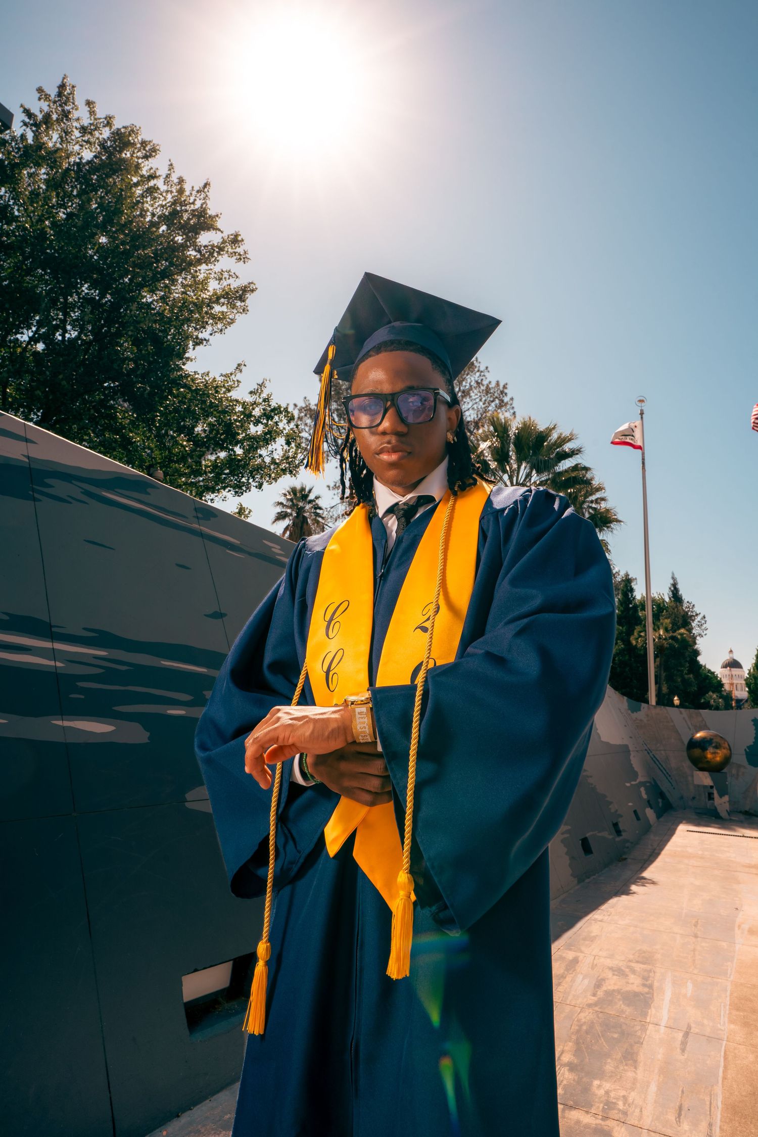 Graduate in blue and gold cap and gown posing outdoors in bright sunlight.