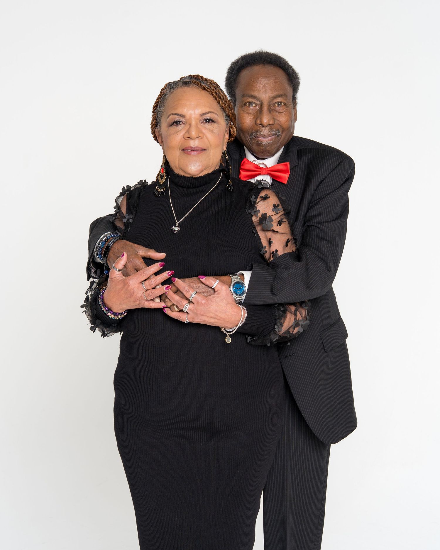 A couple in black formal wear embrace warmly in a professional white background studio portrait.