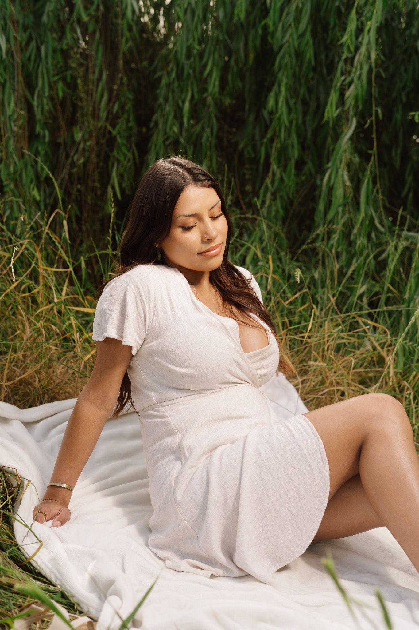 A young woman in a white dress sits peacefully on a blanket surrounded by tall green foliage.
