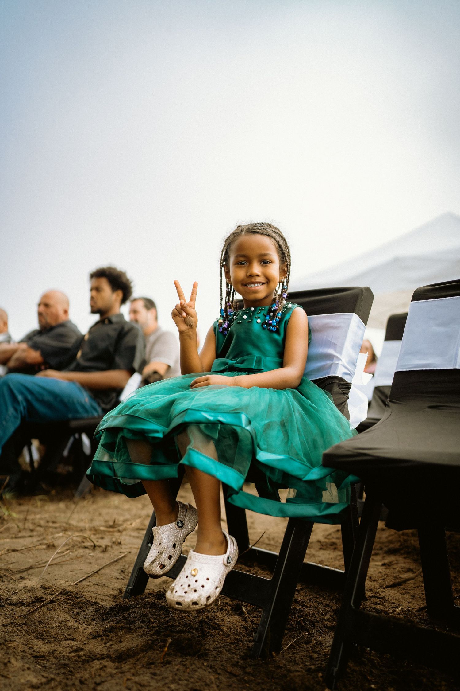 Adorable young girl in green tulle dress flashing peace sign at outdoor wedding ceremony.