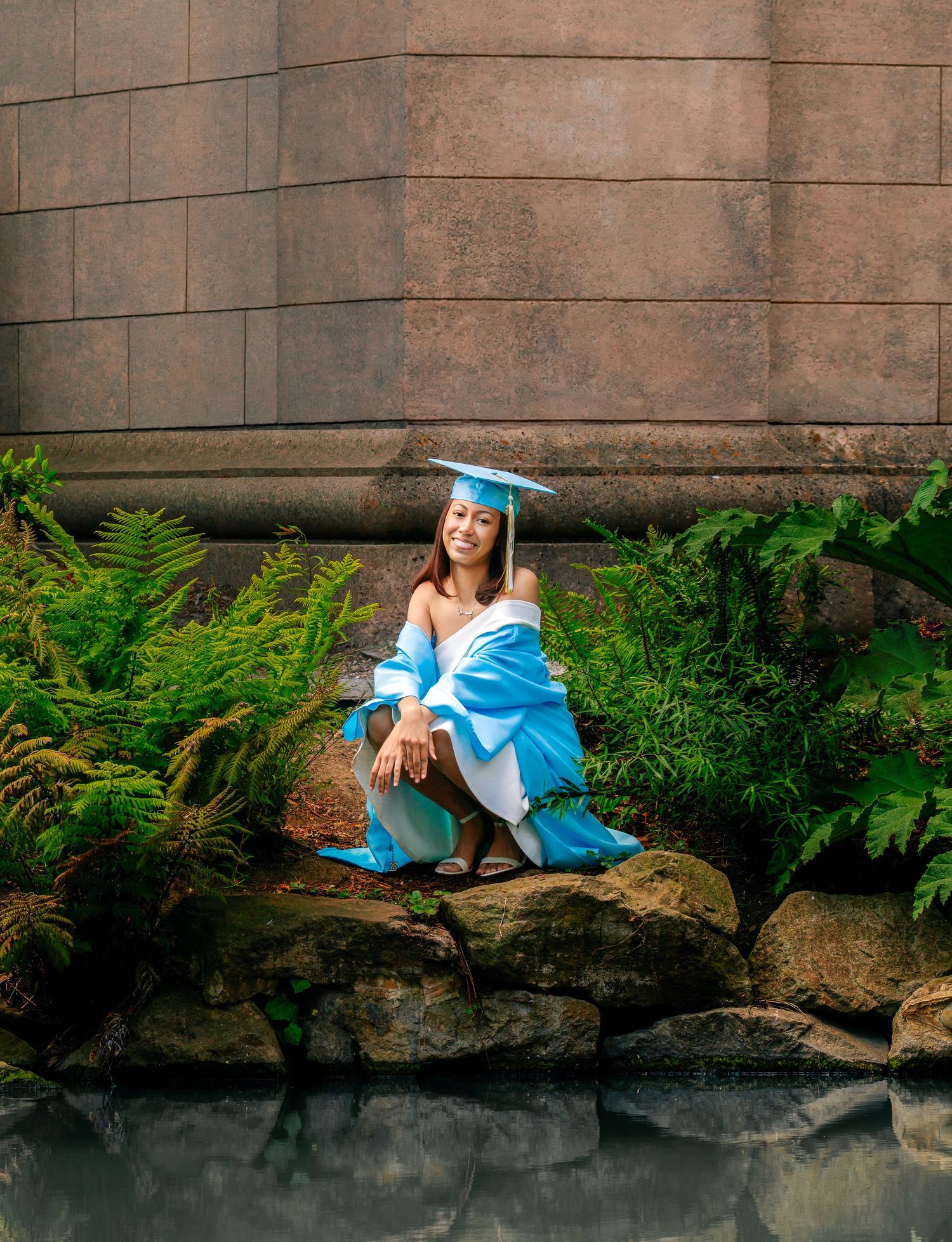 Graduate in blue cap and gown sitting on rocks near a pond surrounded by lush ferns.