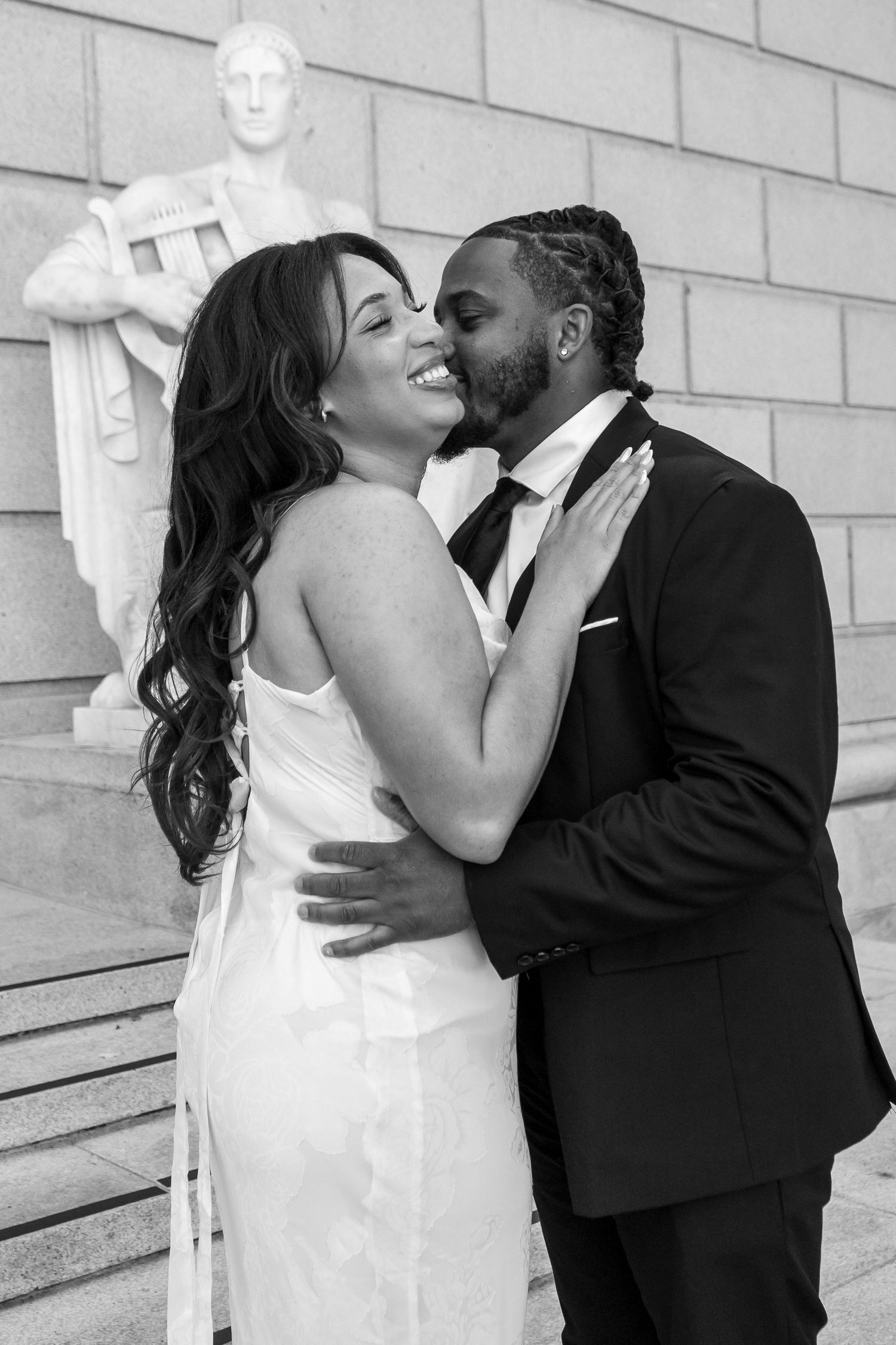 Black and white photo of laughing bride and groom kissing on steps near classical statue.