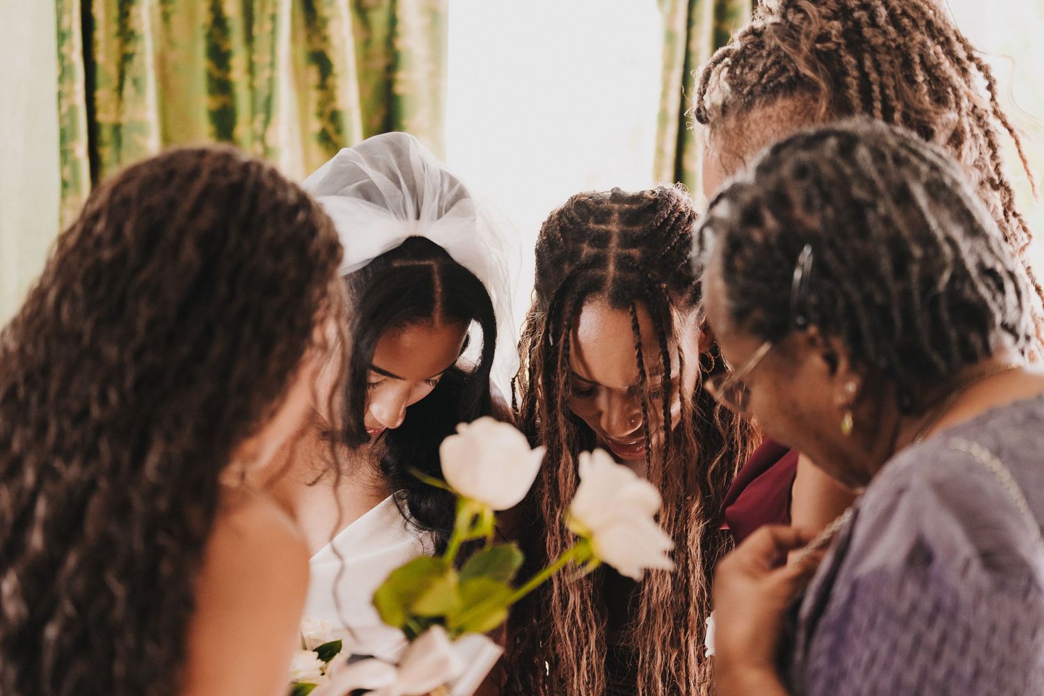 Bride surrounded by family members in intimate prayer circle holding white roses before wedding ceremony.
