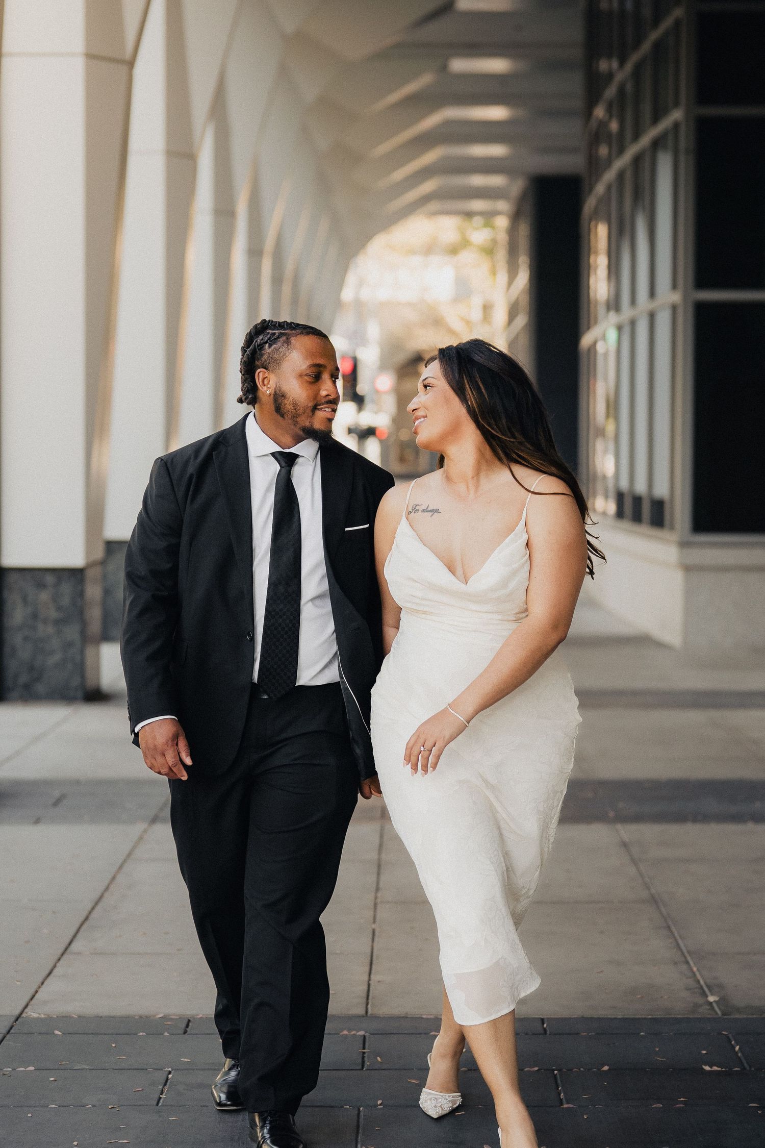Elegant couple walking together, bride in white slip dress and groom in black suit along a city colonnade.
