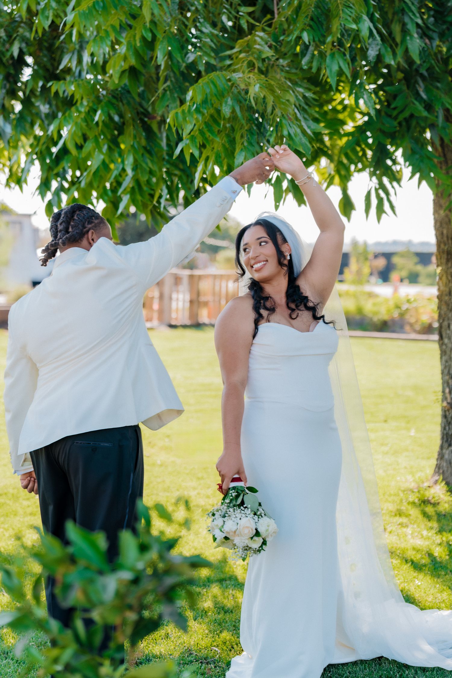 Groom twirls smiling bride in white dress under green tree on sunny wedding day.