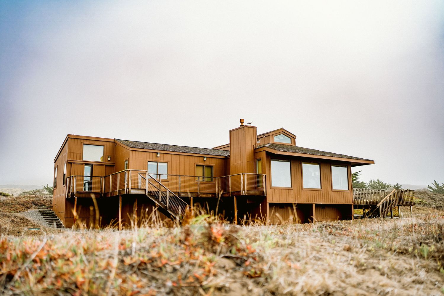 Rustic brown coastal vacation home with large deck surrounded by golden grass under overcast sky.