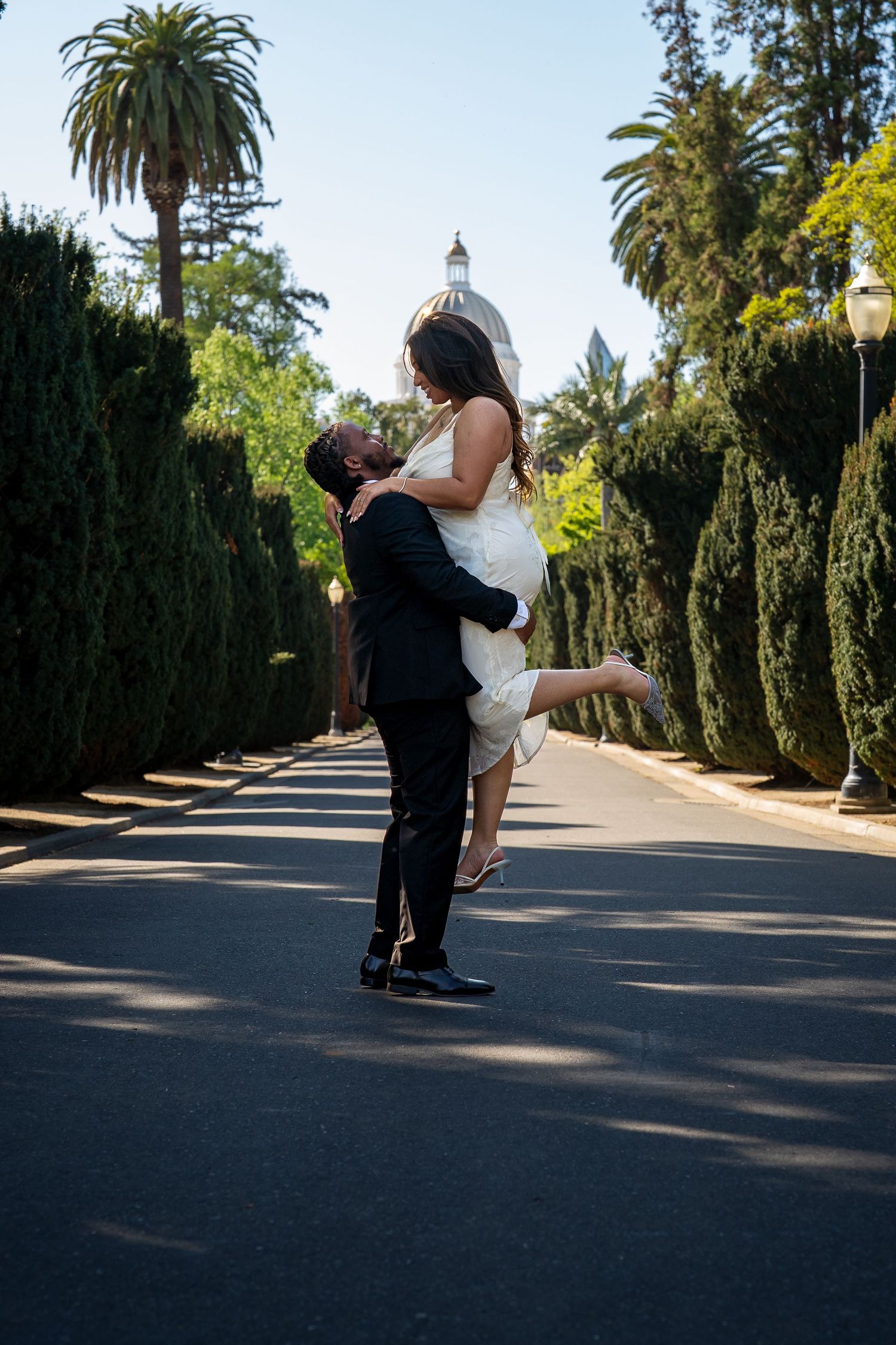 Groom lifts bride in white dress on tree-lined avenue with domed building backdrop on wedding day.