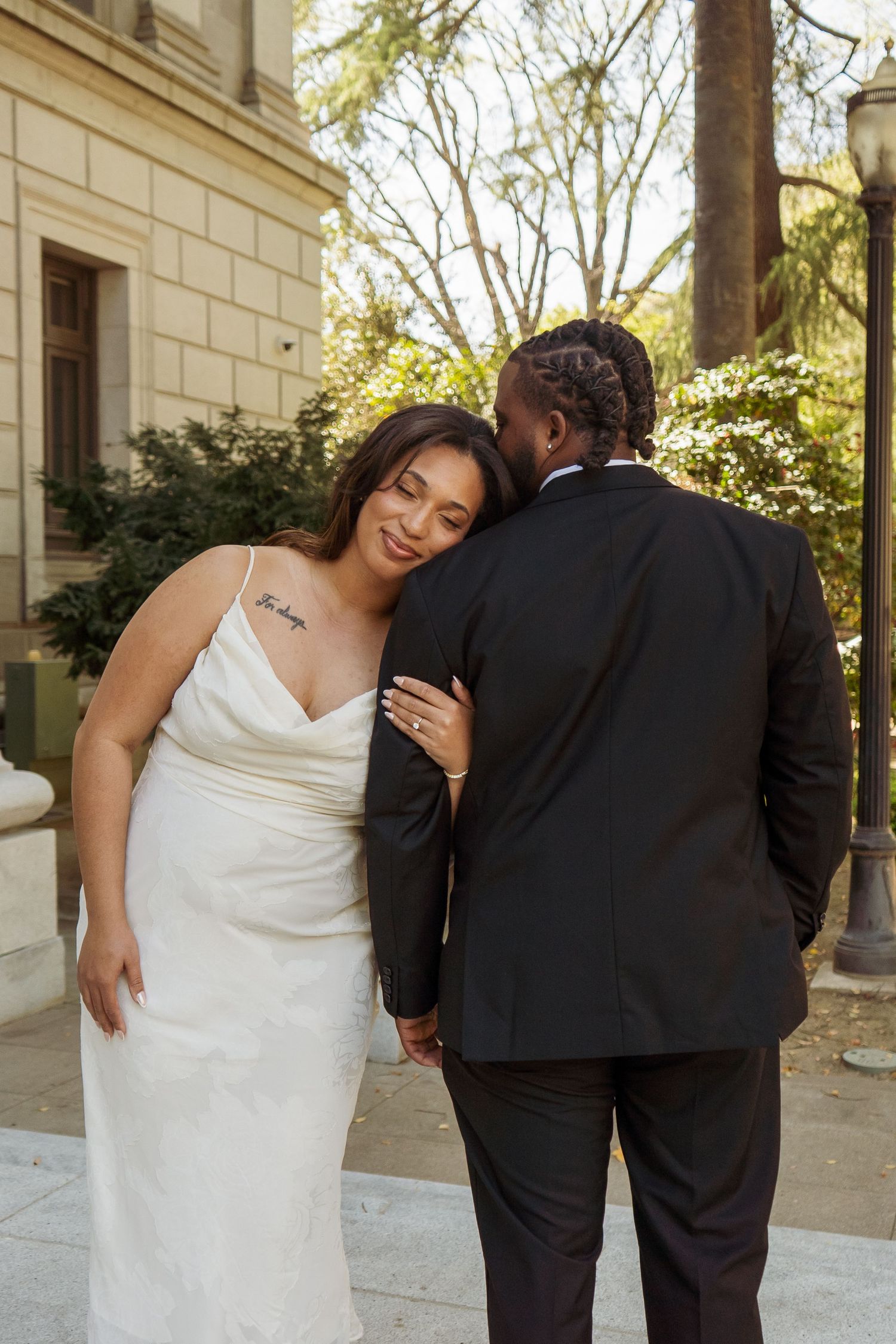 Bride in white satin gown rests head on grooms shoulder during outdoor wedding portraits.