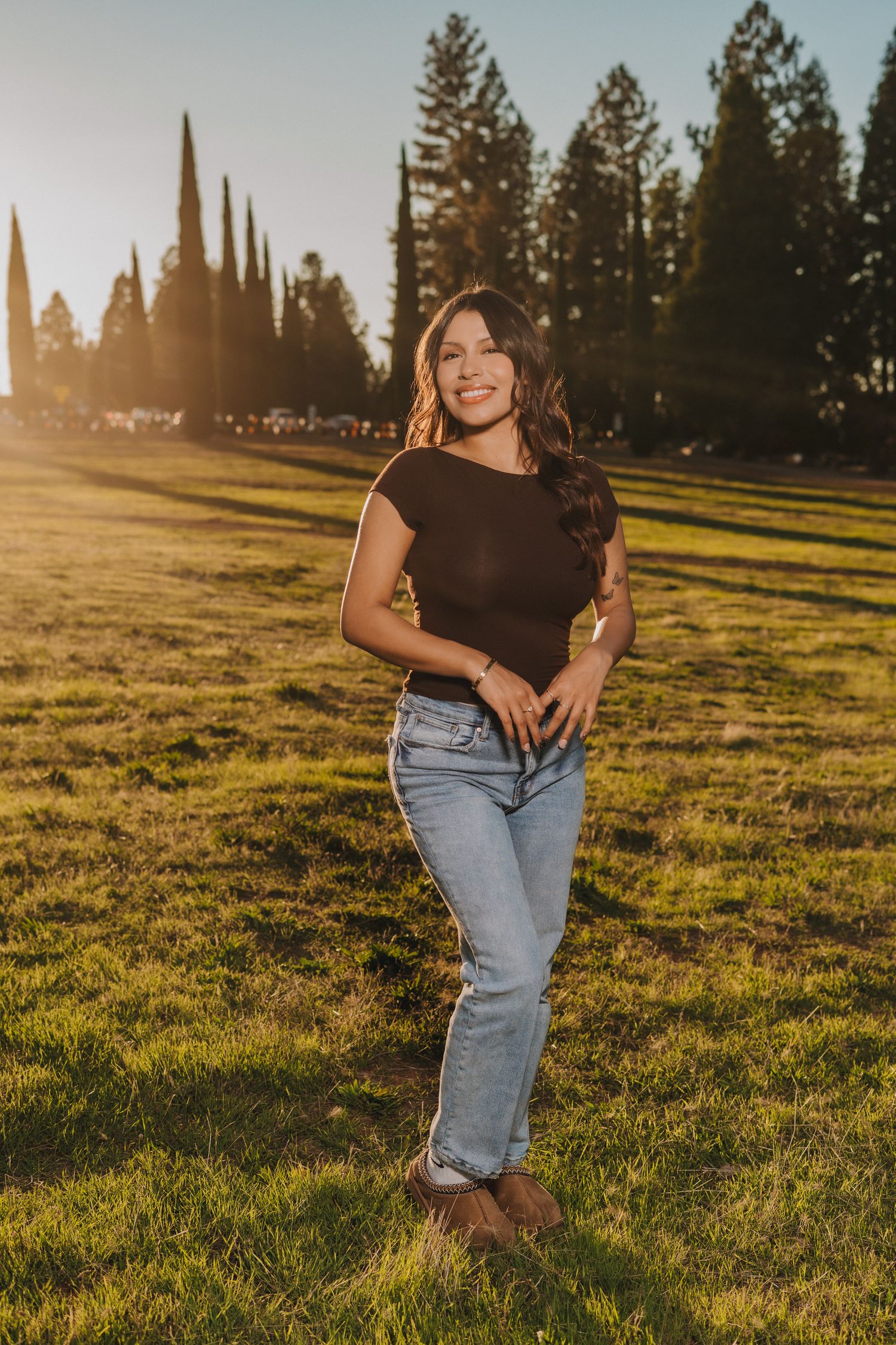 Young woman in black top and jeans smiles in a sunlit park with tall trees at golden hour.