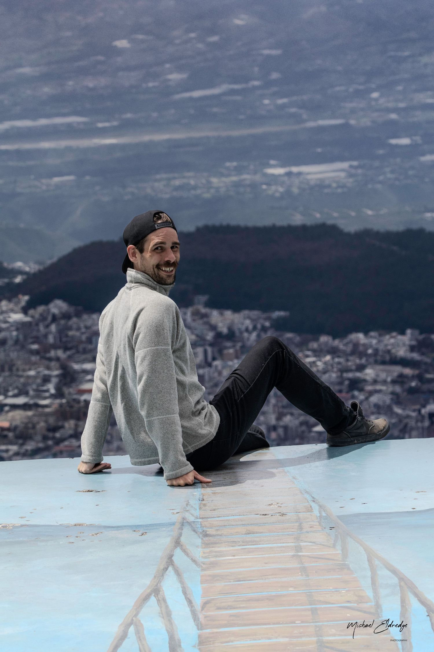 Person relaxing on sloped rooftop with cityscape and mountains visible in background.