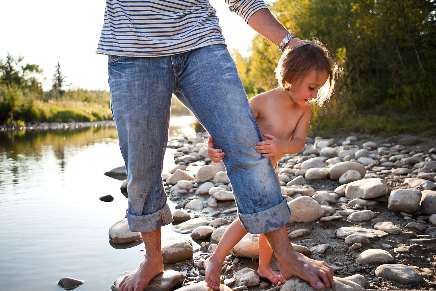 Parent holding a toddler's hands as they play together on rocks near a river at golden hour