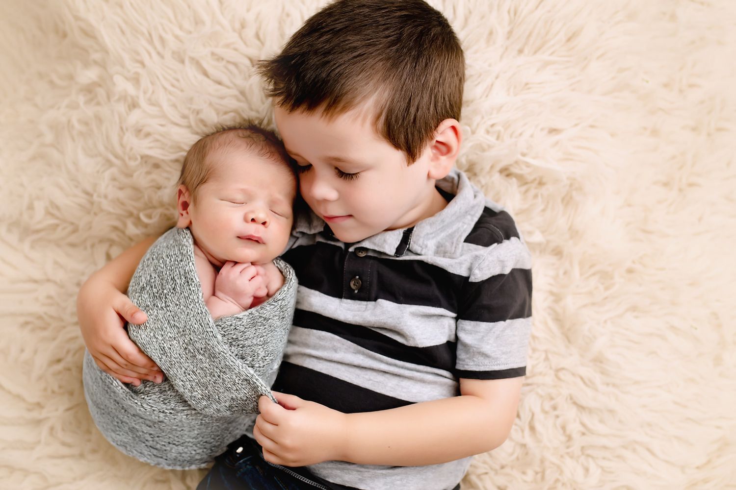 Young boy gently cradling his sleeping newborn sibling wrapped in a grey wrap on a white fluffy blanket