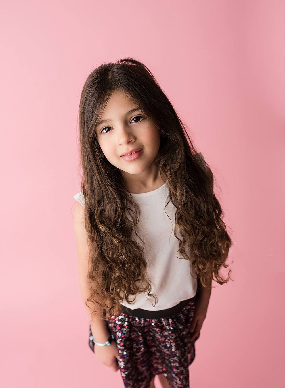 Young girl with curly brown hair wearing a stylish outfit against a pink studio background