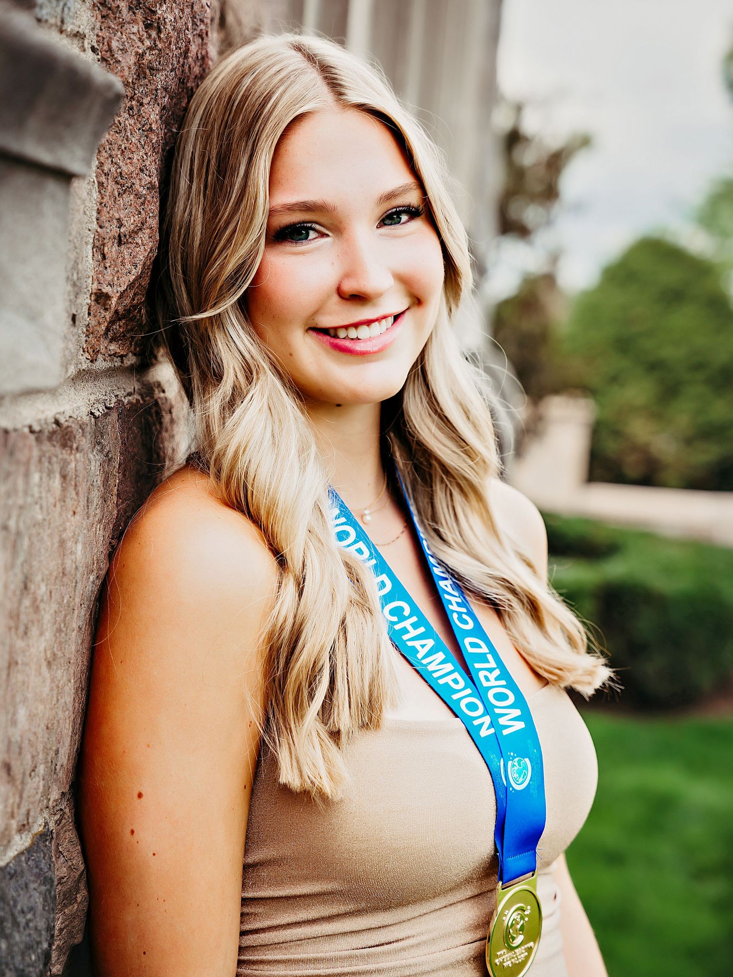 Young blonde woman wearing a gold medal smiling against a stone wall in an outdoor portrait