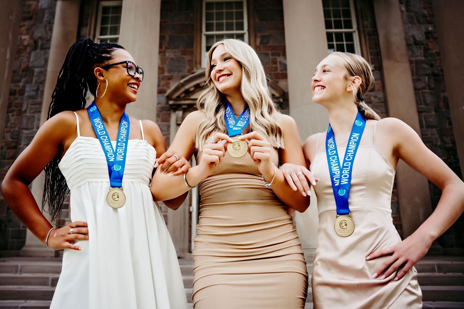 Two young women wearing gold medals laughing together in front of a historic stone building