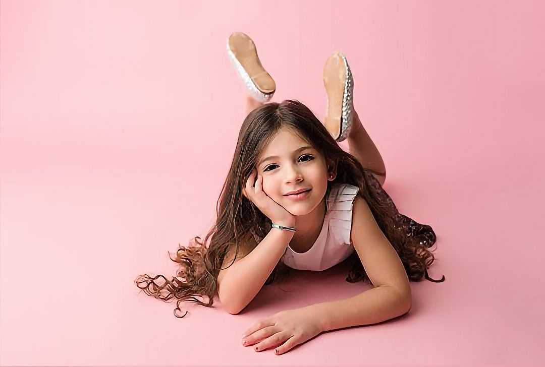 Young girl with long dark hair posing with chin on hands in a pink studio portrait