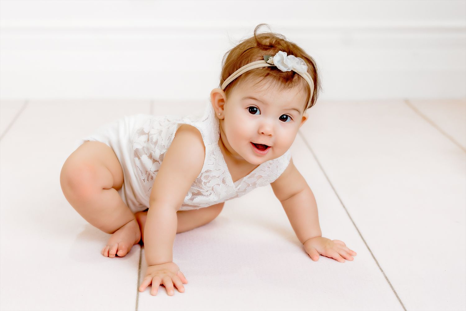 Baby crawling on a white studio backdrop wearing a white outfit and delicate headband