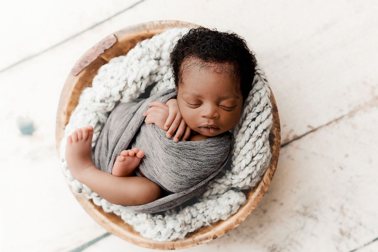 Sleeping newborn baby wrapped in a grey swaddle nestled in a wooden bowl prop on a white background